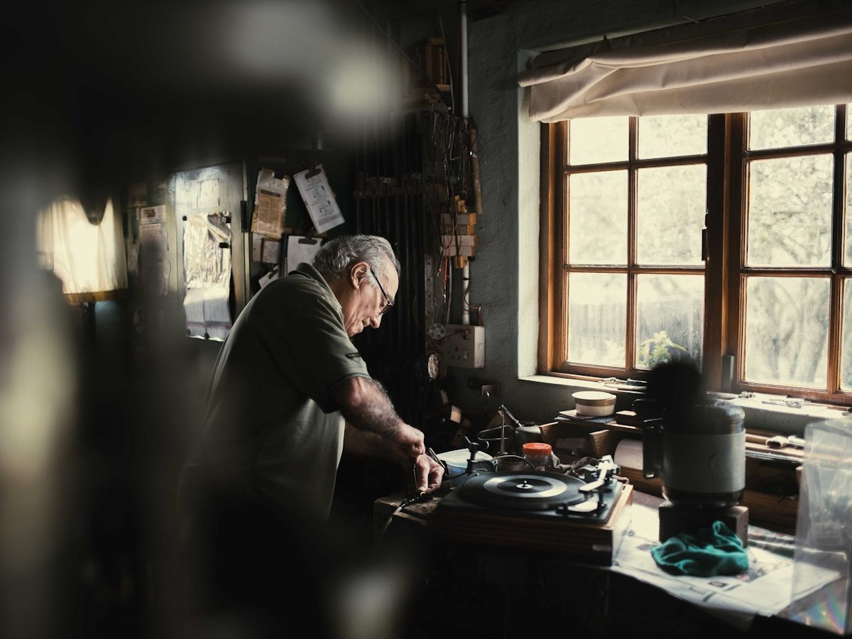 A man working with hand tools at a lamp-lit workbench beside a window